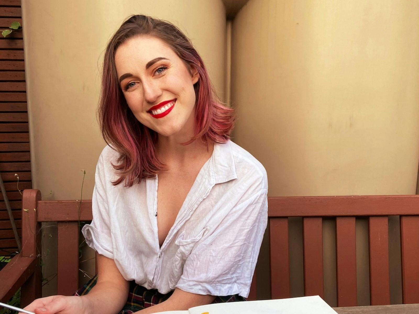 A smiling woman with brown hair with pink highlights sits on a wooden bench outdoors.