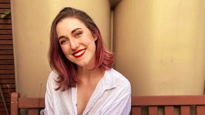 A smiling woman with brown hair with pink highlights sits on a wooden bench outdoors.