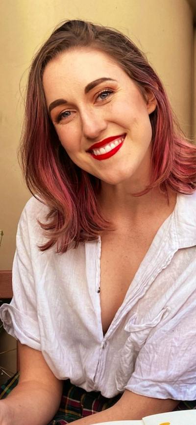 A smiling woman with brown hair with pink highlights sits on a wooden bench outdoors.