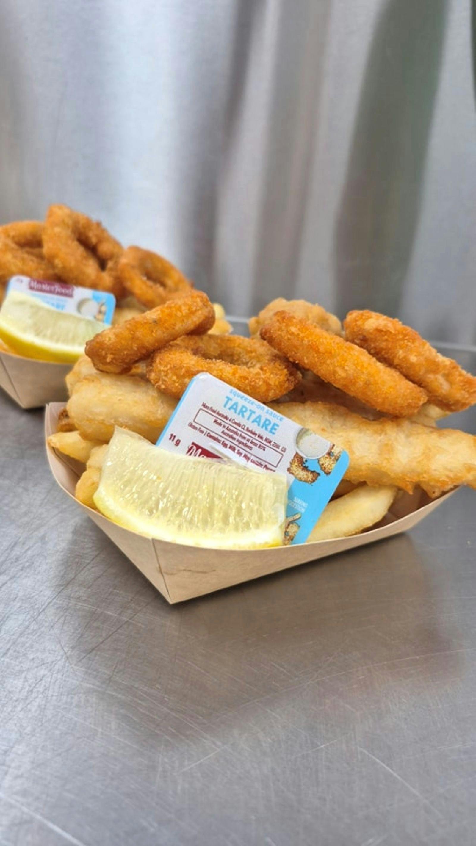 Paper bowl with calamari rings, fish and chips with tartar sauce and a slice of lemon.