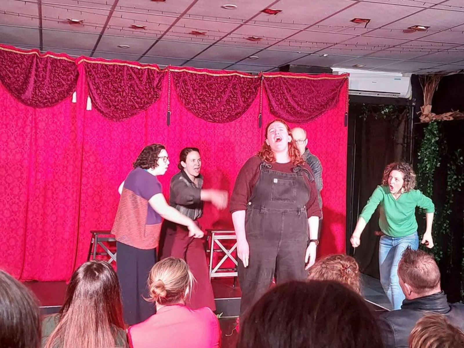 5 People standing singing on a stage with dramatic actions. Audience watching.