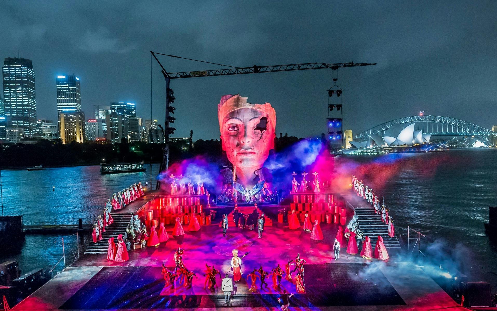 Aida performers fill a waterfront stage beneath a giant Nefertiti face, with Sydney Harbour behind