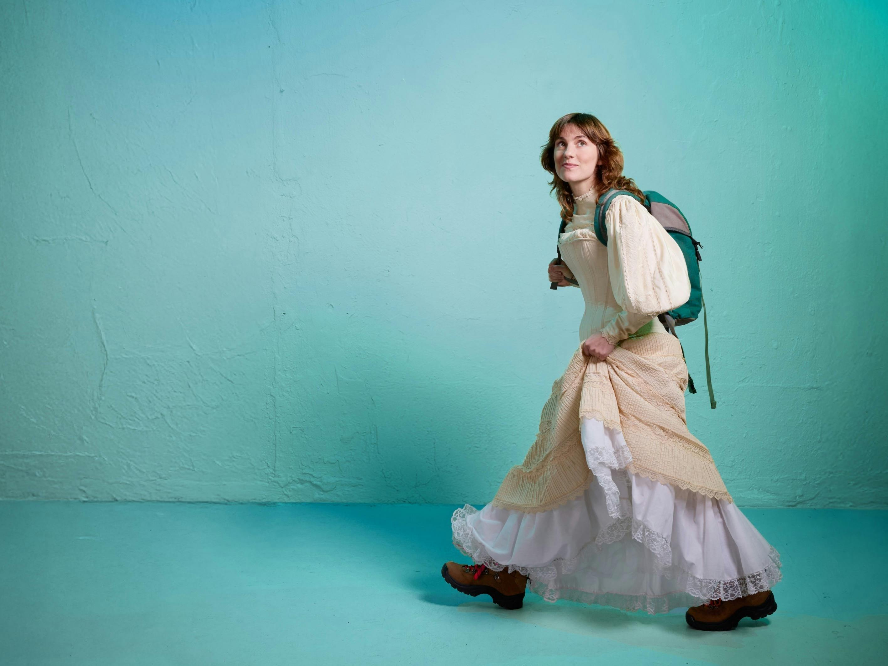 A young woman in a flowing cream dress walks across a turquoise wall with a backpack