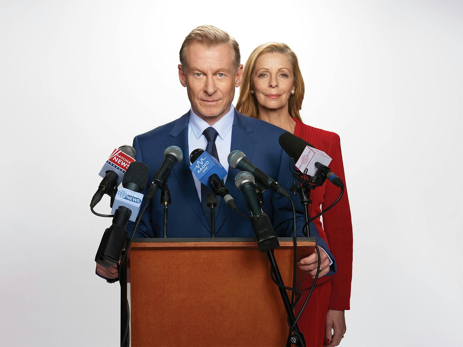 Two people in formal wear stand at a podium with news microphones attached. They look serious.