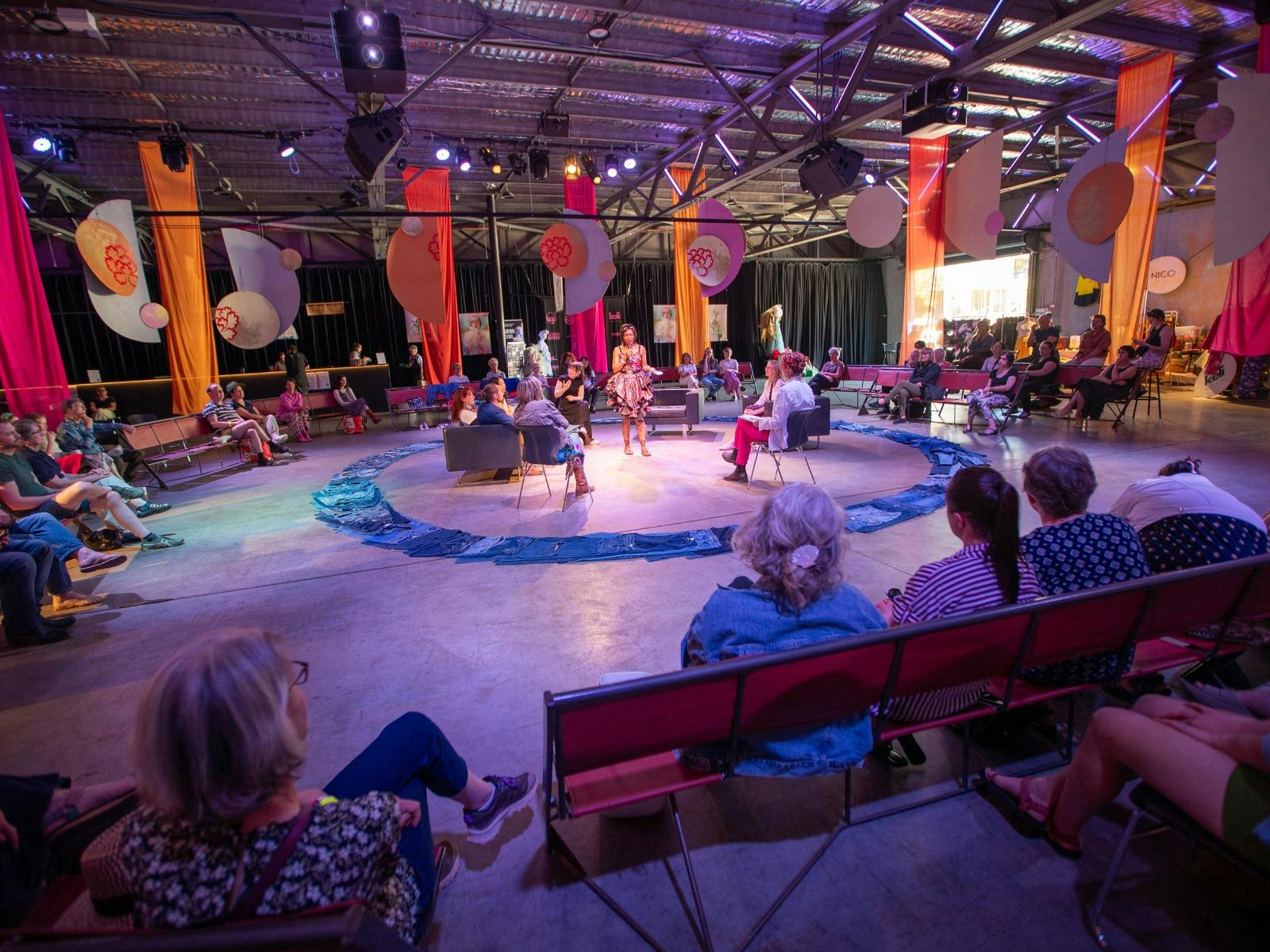 A large space with people sitting around the edges, in the middle, panelists sit in chairs and talk.