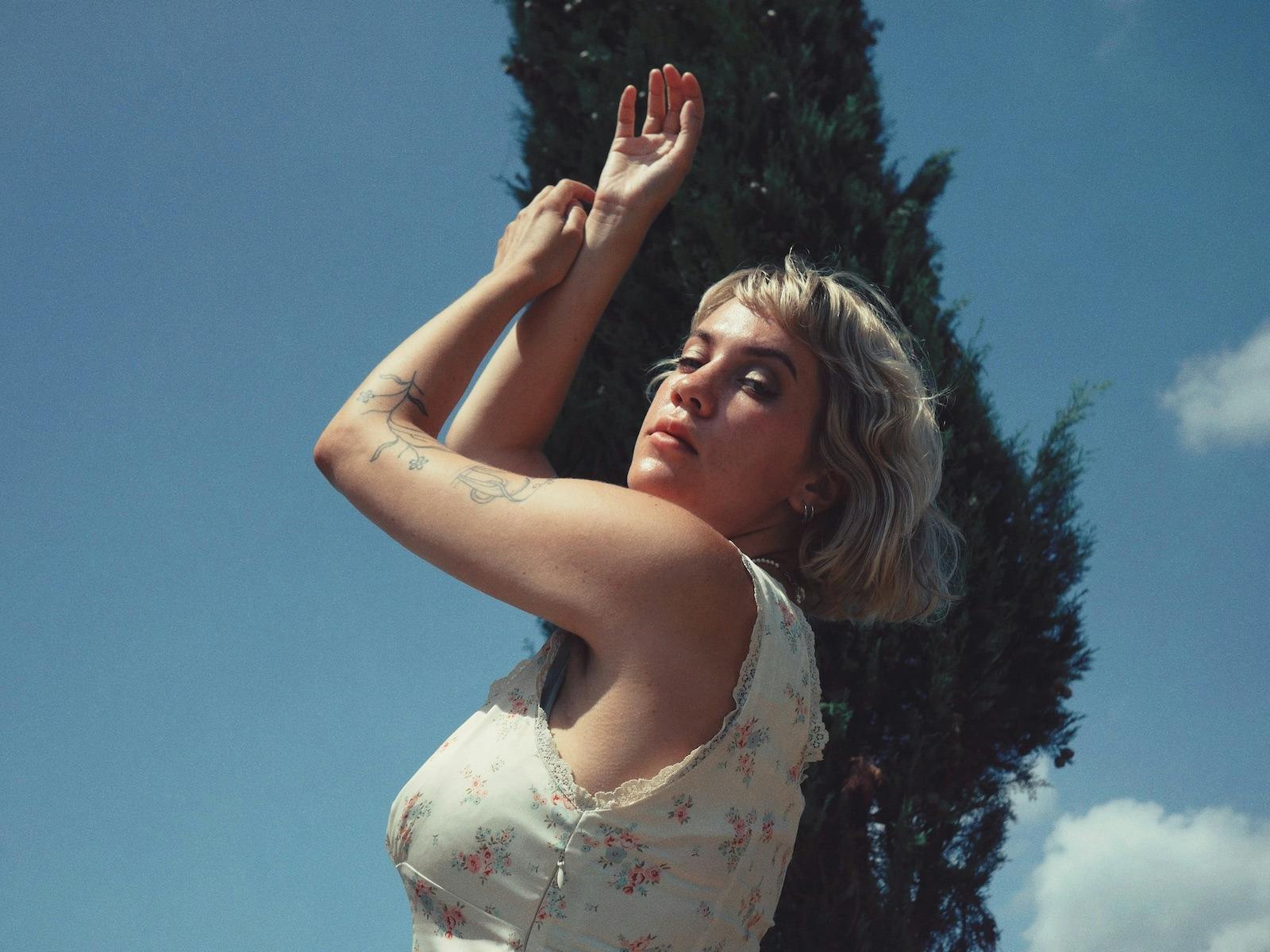 Bess Atwell in front of a blue sky and tall tree with her eyes closed and hands up to the sky.
