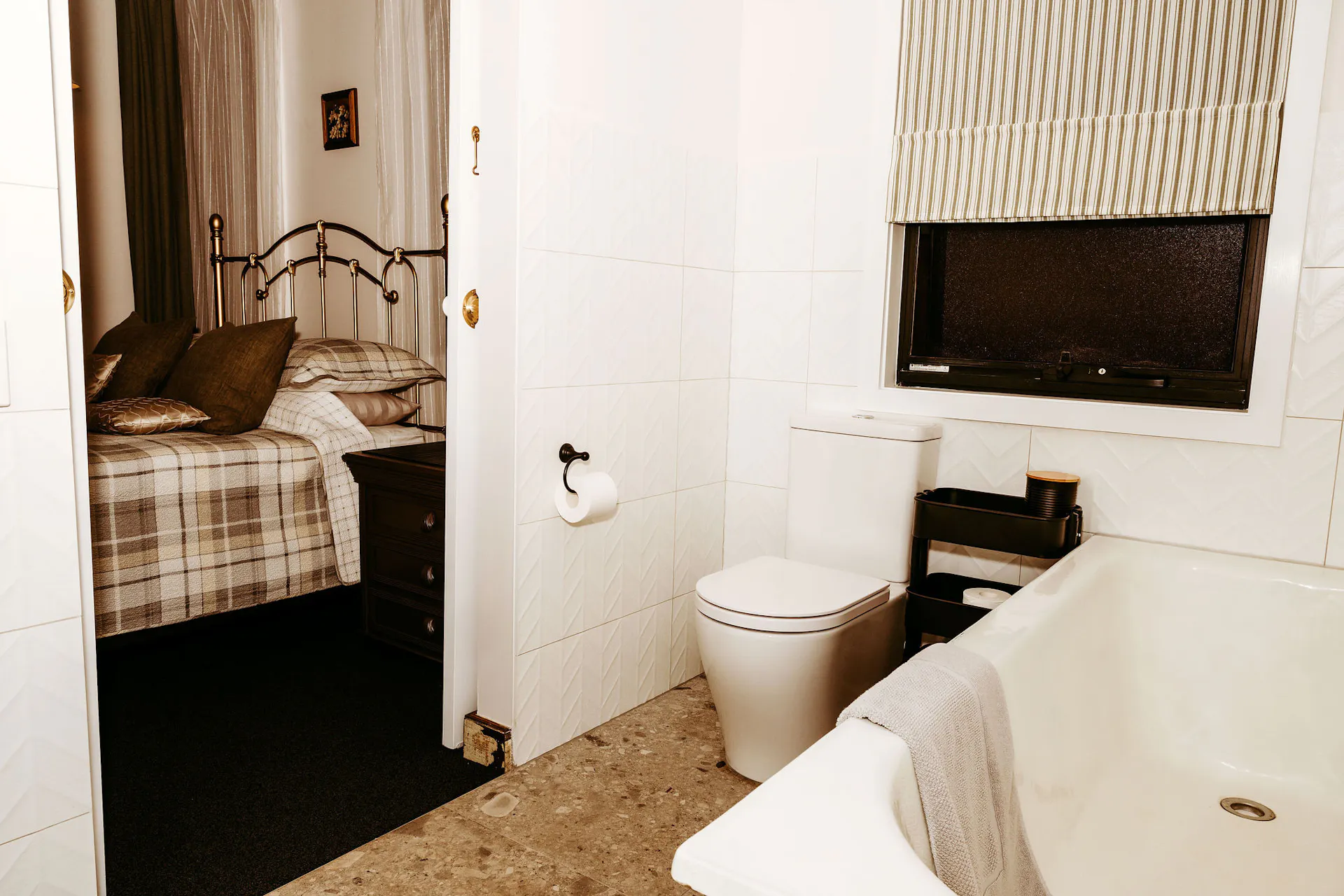 Image taken from ensuite through to bedroom and shows bath tub toilet and bed with check linen