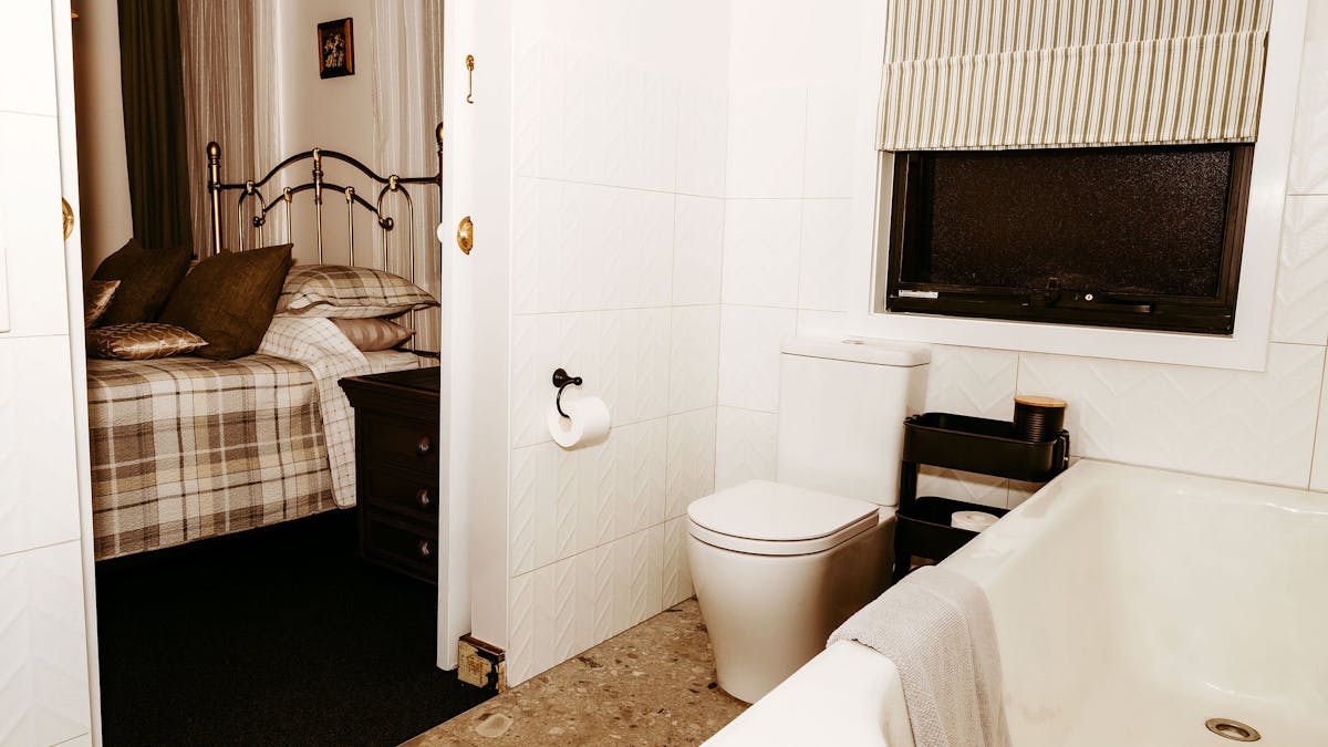 Image taken from ensuite through to bedroom and shows bath tub toilet and bed with check linen