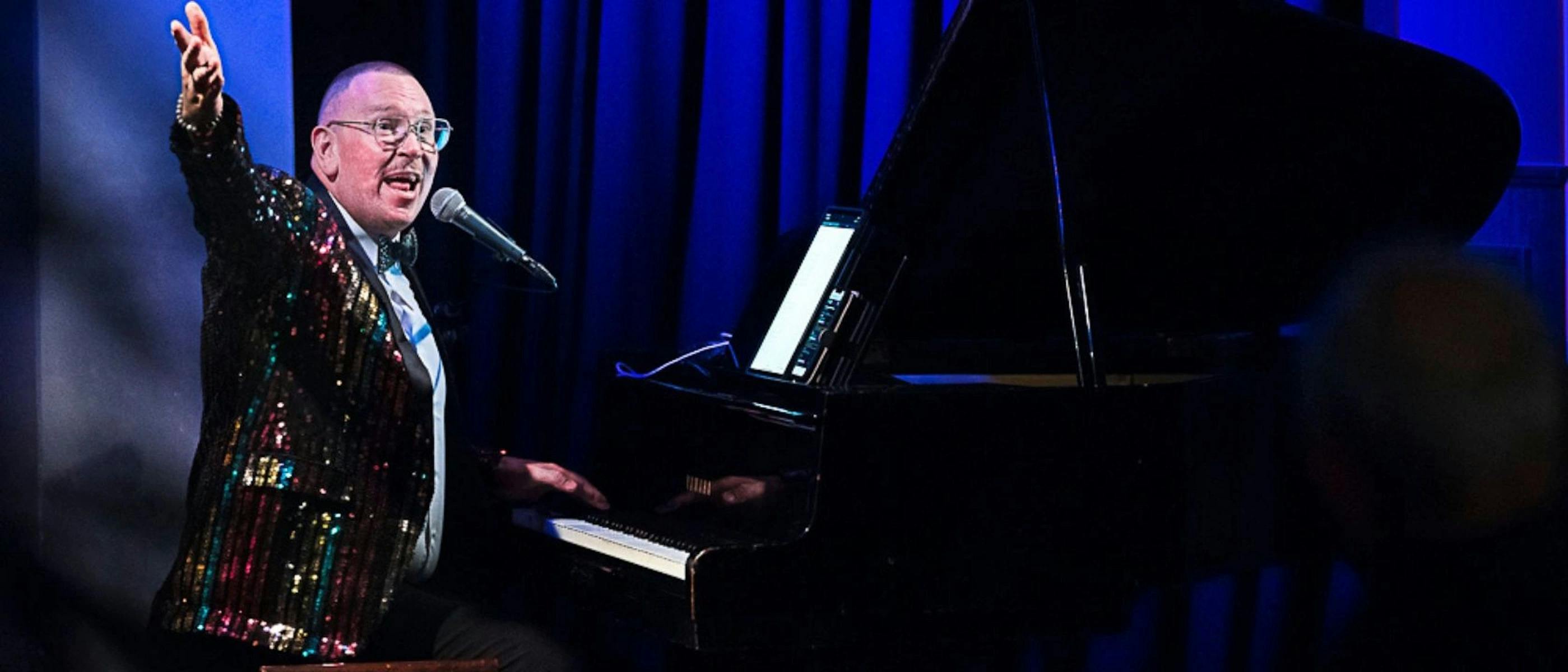 A man wearing a rainbow striped jacket sitting at a piano, raising his arm.