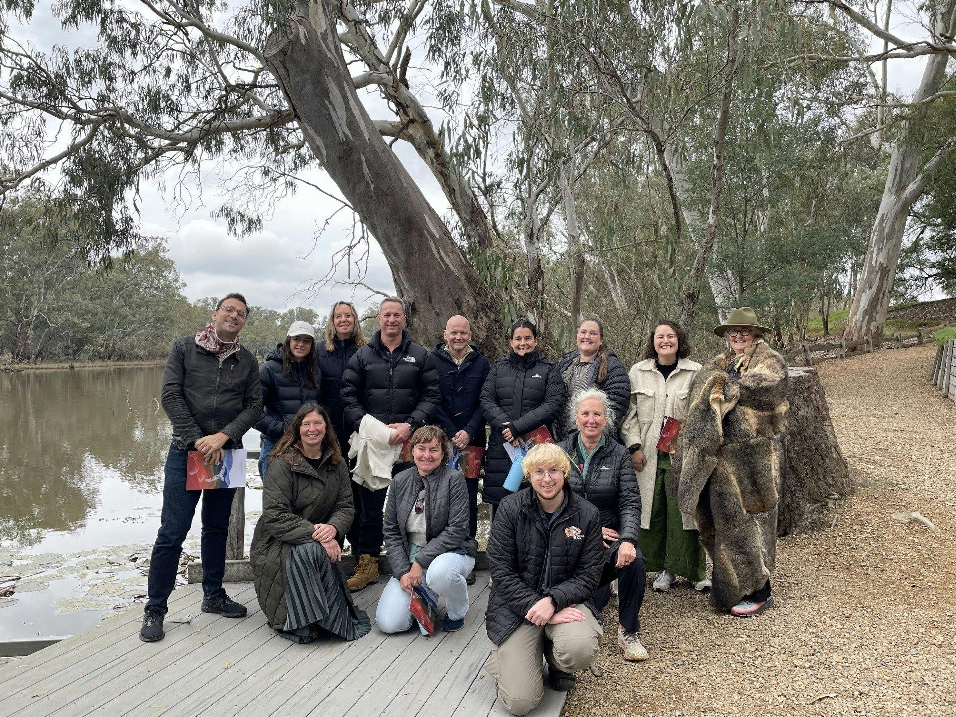 a group of people facing the camera standing near a river around trees