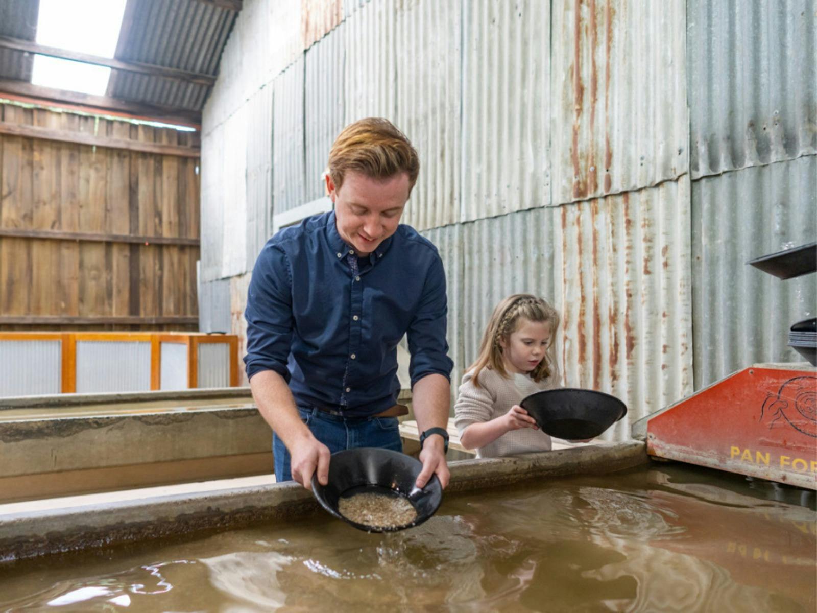 A young father is scooping sand with a gold pan. His daughter looking at her gold pan to spot gold.