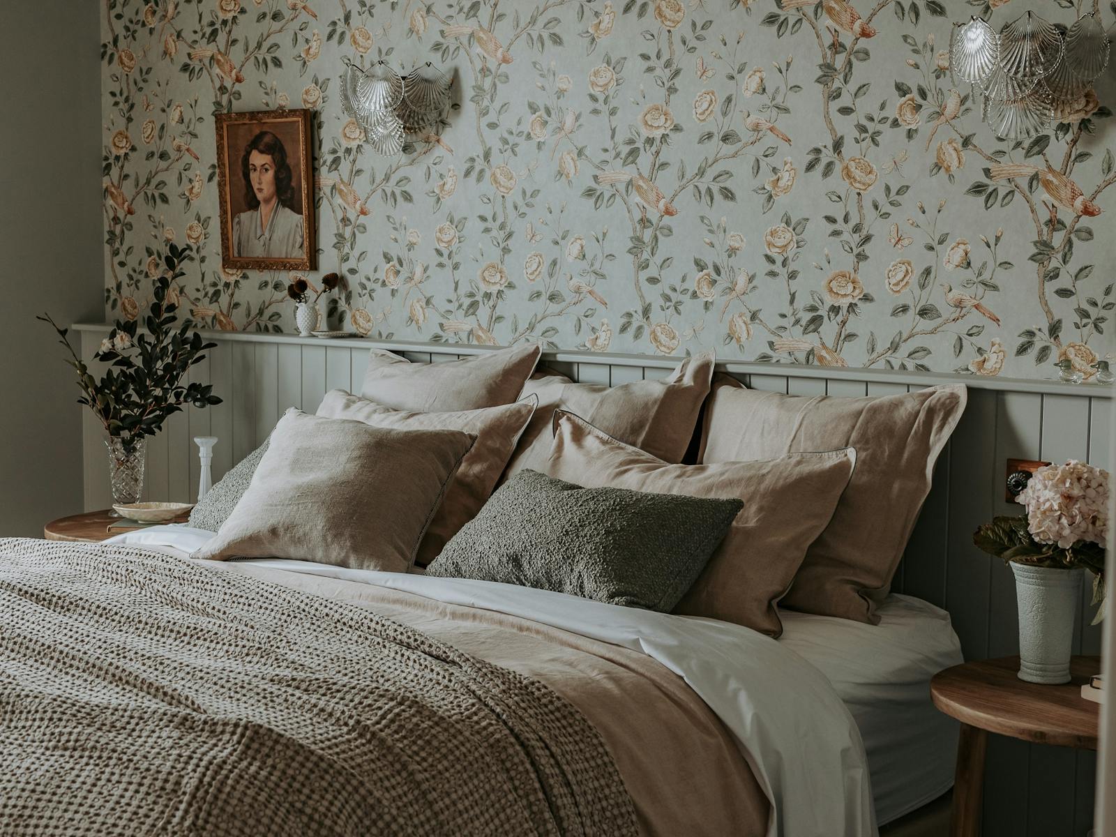 King size bed, floral wallpaper, wall sconces, artwork and vase of greenery