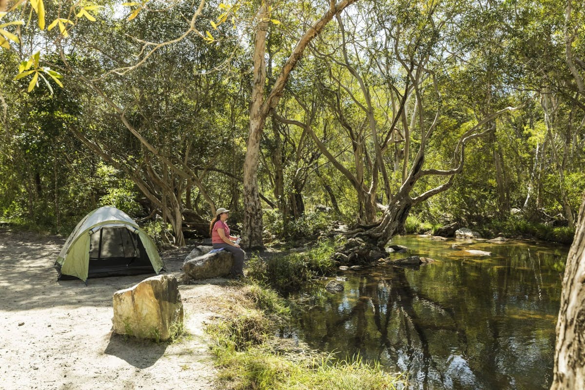 Person sitting on rock looking at creek with a tent set up behind them.