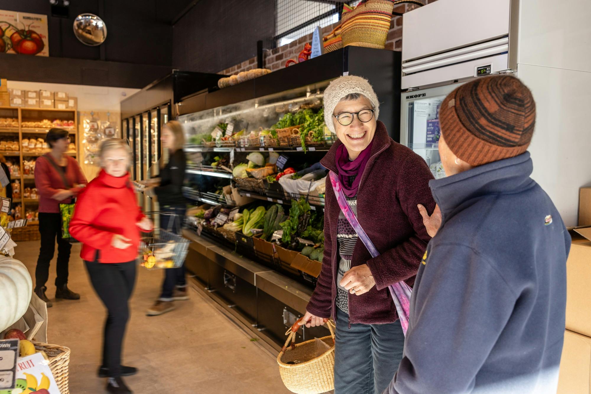 Woman smiles as another woman touches her on the shoulder, fresh veggies in a display case behind