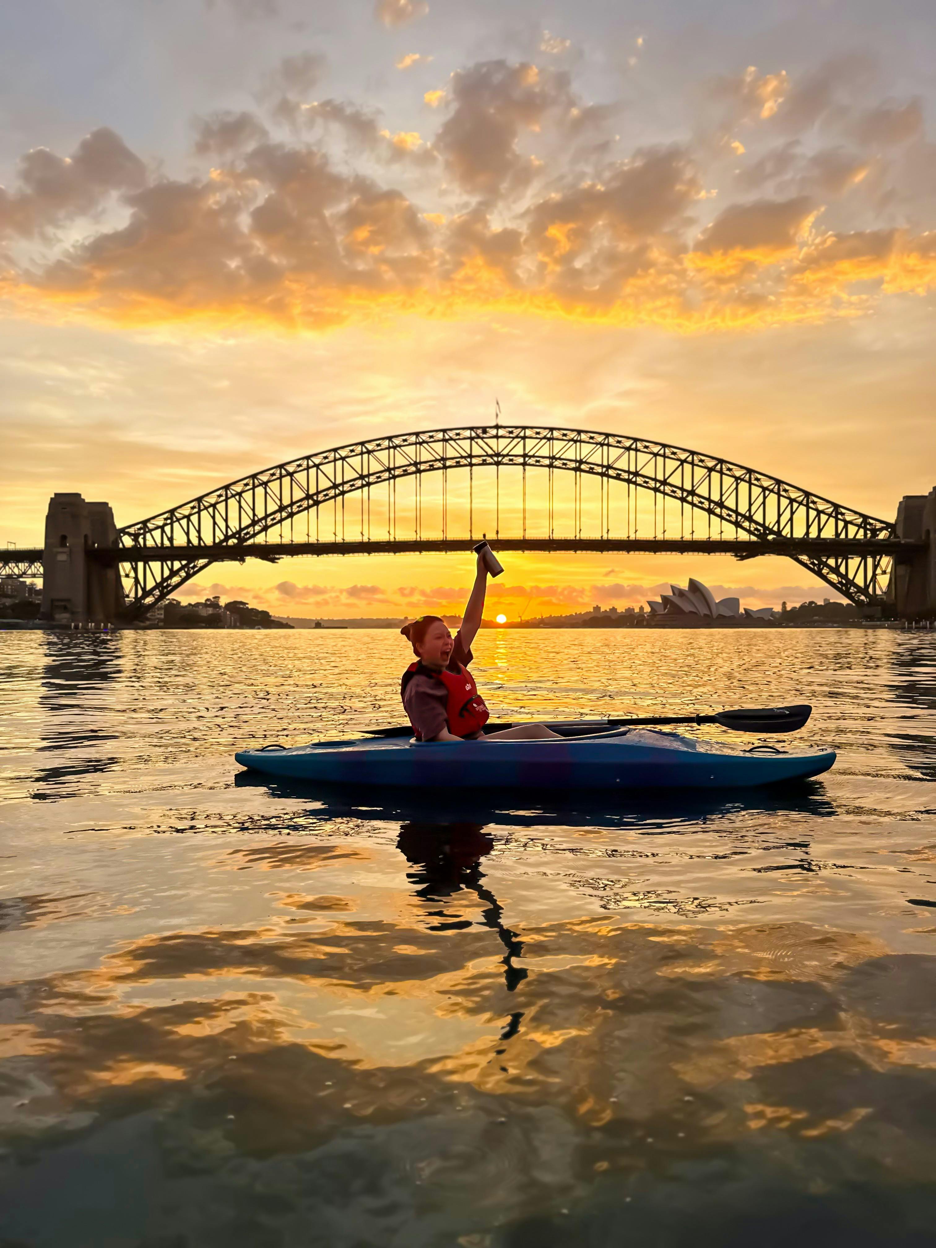 Kajakfahrer hält Kaffee hoch in den Himmel, im Hintergrund das Opernhaus und die Sydney Harbour Bridge.