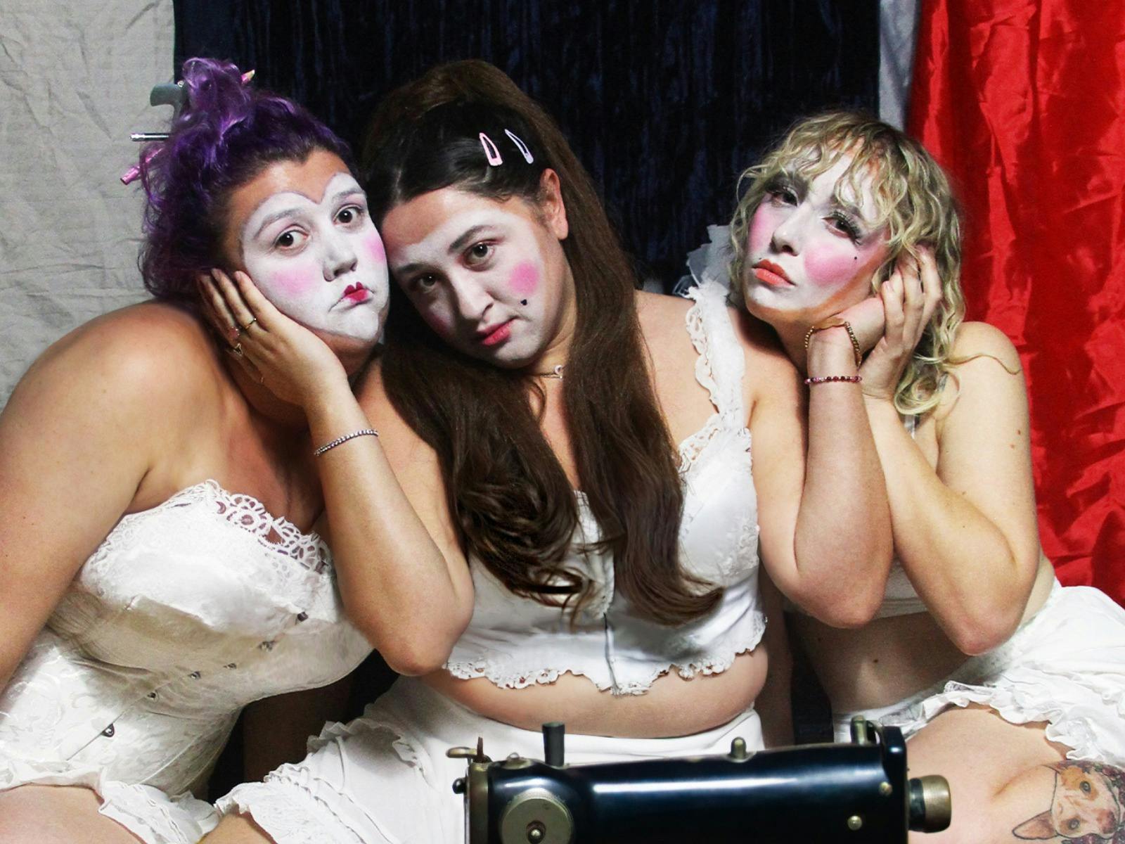 3 young women in white corsets, bloomers and clown makeup lean on each other surrounded by material.