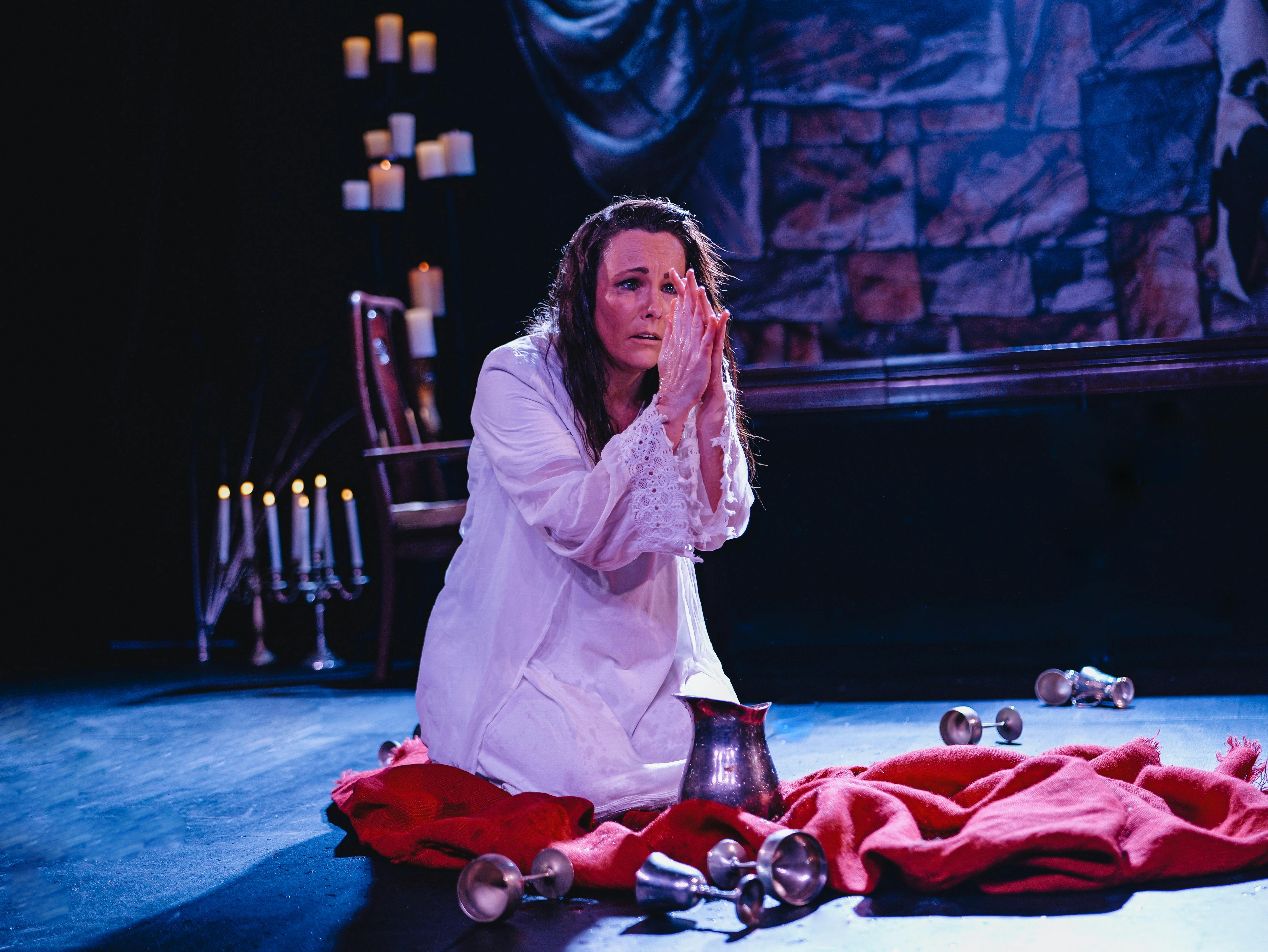A woman in a white dress kneels in a praying position surrounded by cups and red fabric
