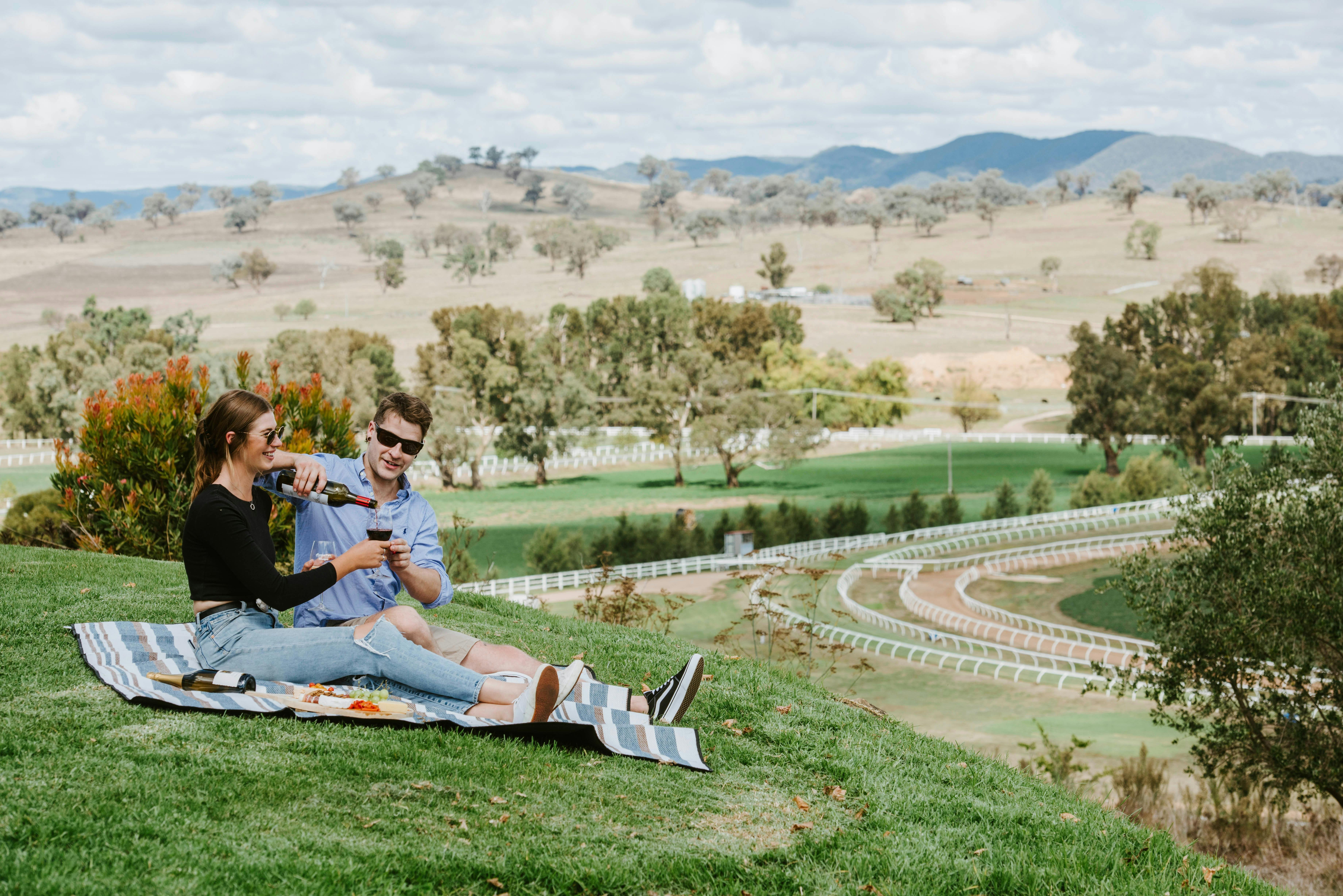 A couple drinking wine on a picnic blanket on a grassy hill above a horseracing track at Gooree Park