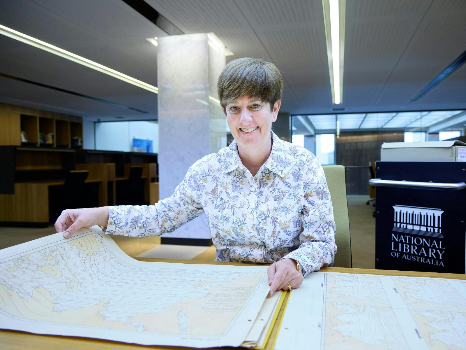 A smiling woman in a floral shirt sits at a desk, holding open a large historical map.