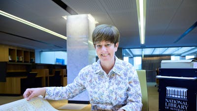 A smiling woman in a floral shirt sits at a desk, holding open a large historical map.