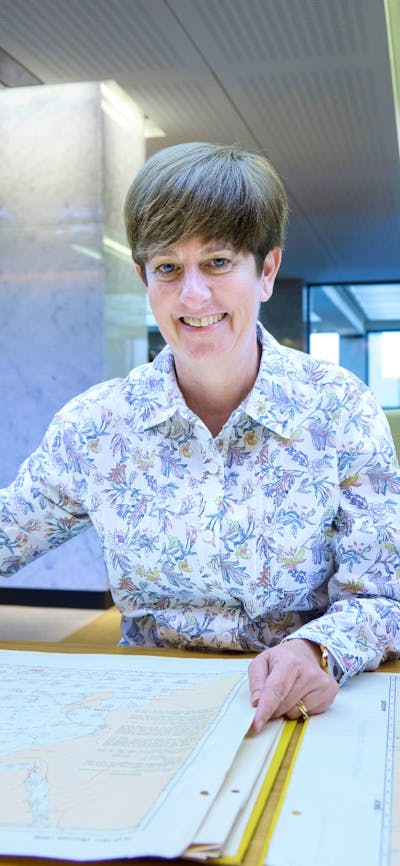 A smiling woman in a floral shirt sits at a desk, holding open a large historical map.