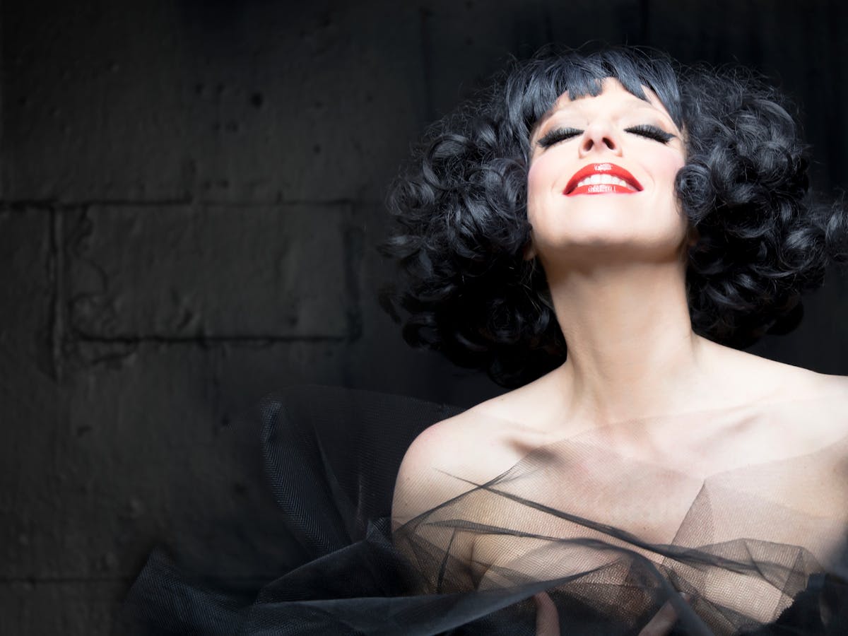 A woman with black curly hair and striking red lipstick, smiling softly against a grey background.