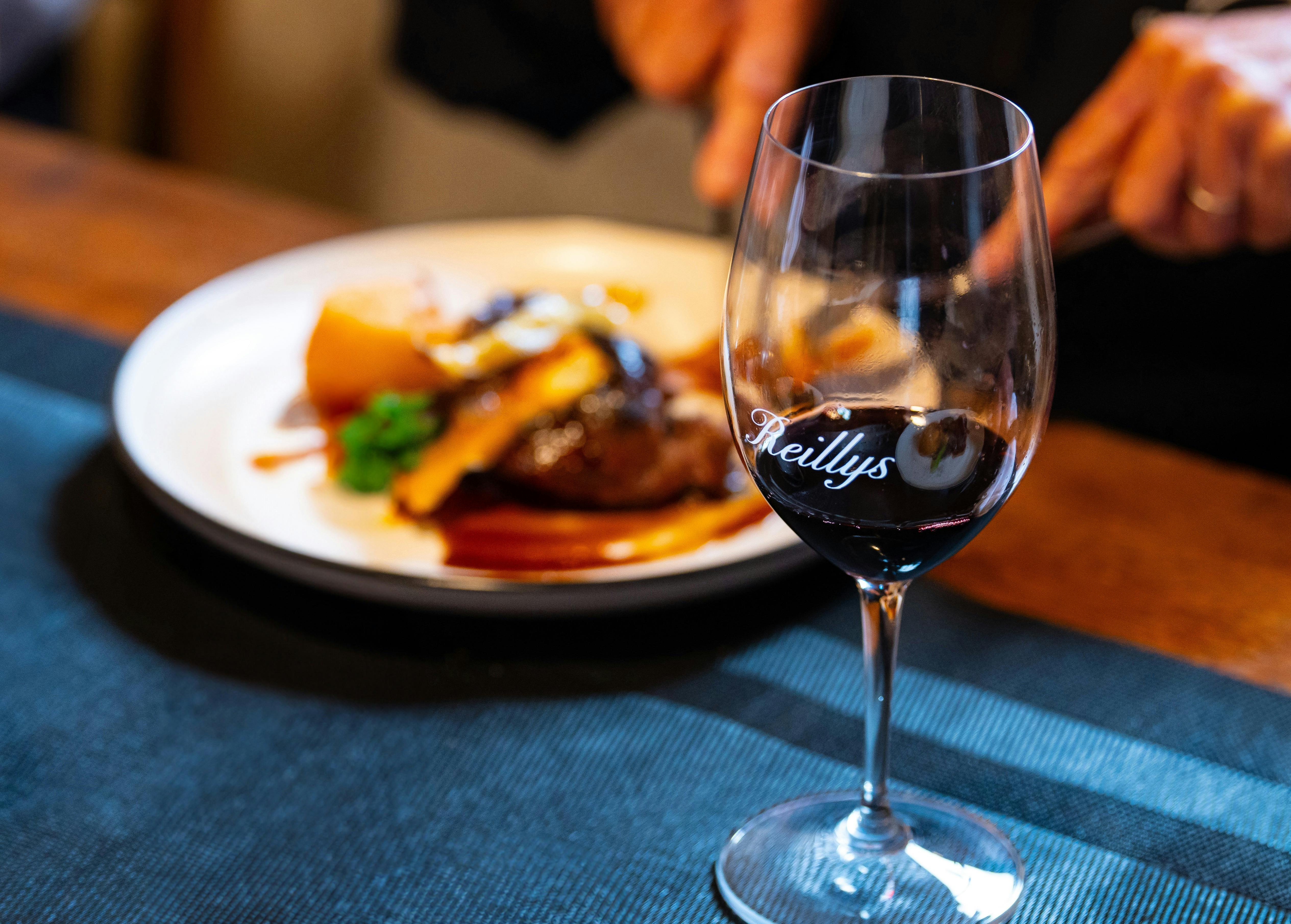 Plate of food out of focus behind a glass of red wine with the Reillys logo on glass