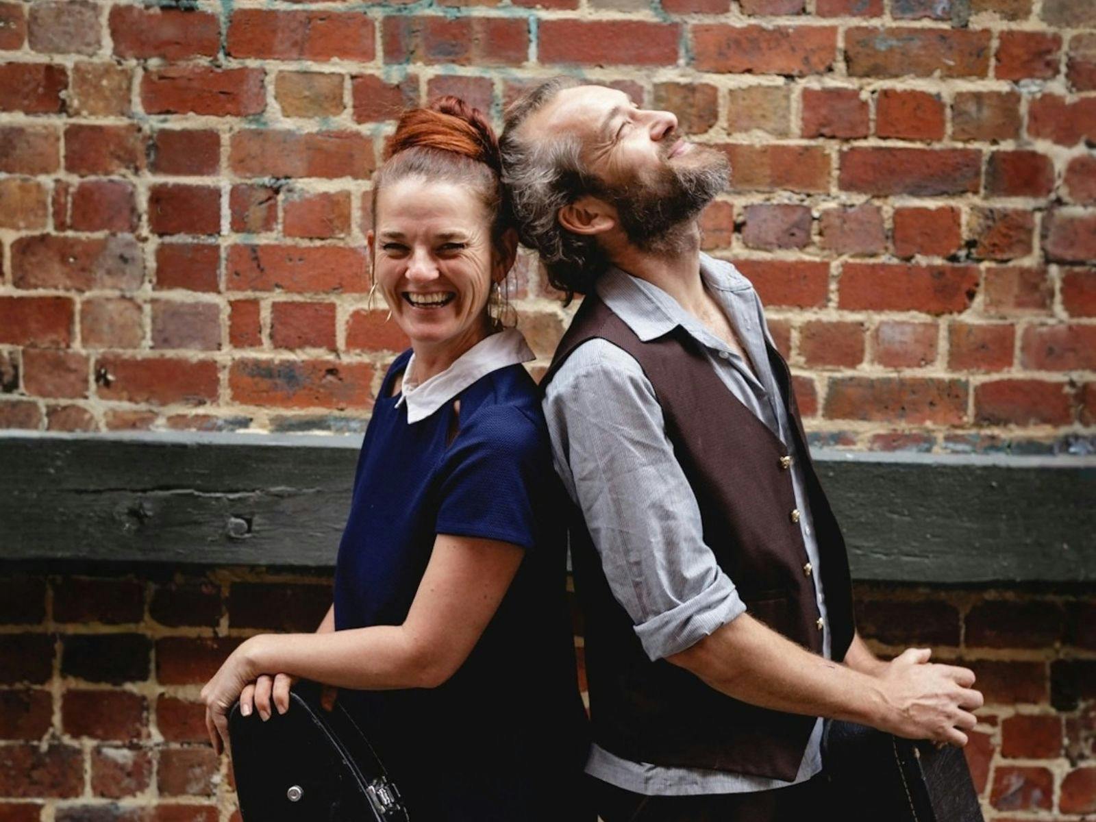 A male and female stand back to back in front of a brick wall laughing.