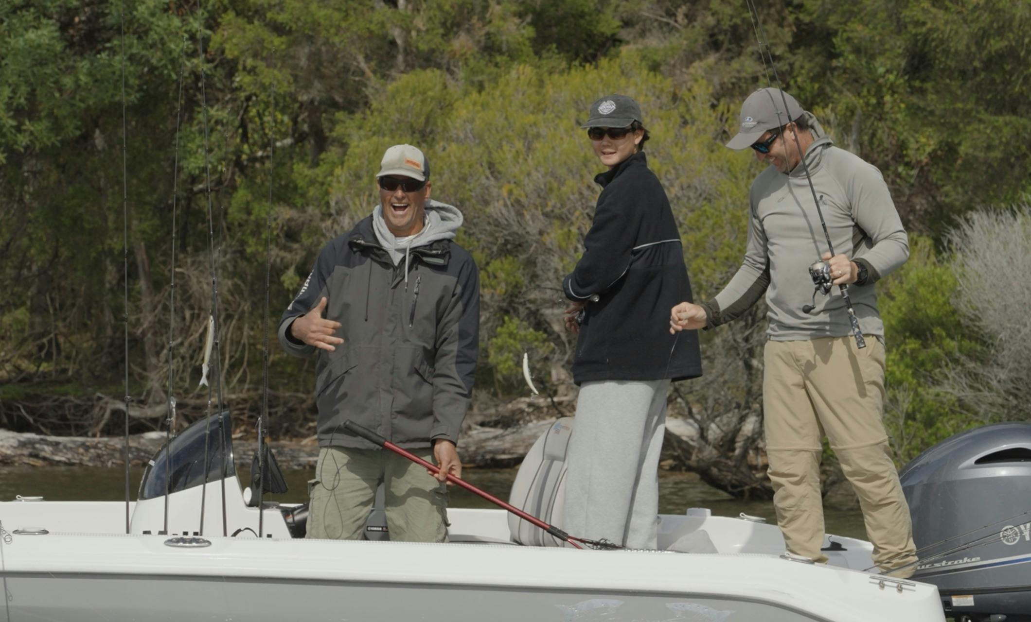 Three anglers laughing and fishing from a boat with dense bushland behind them