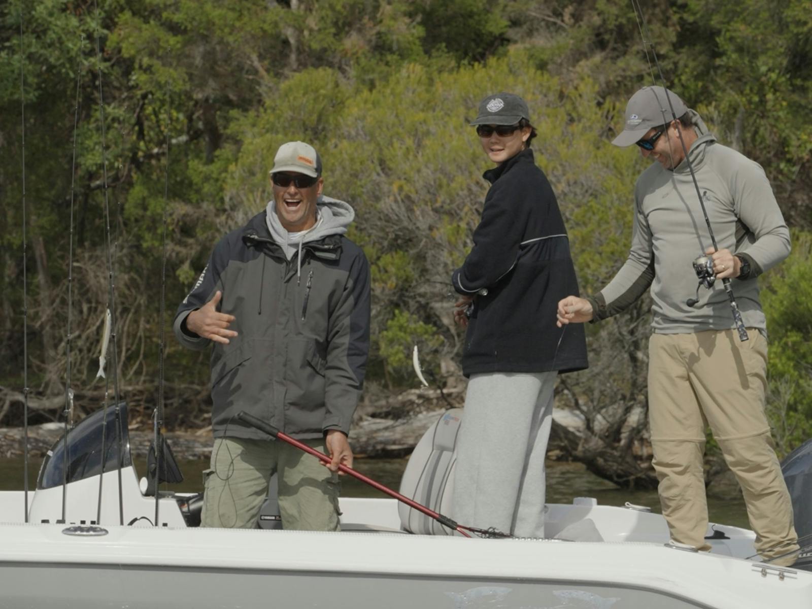 Three anglers laughing and fishing from a boat with dense bushland behind them