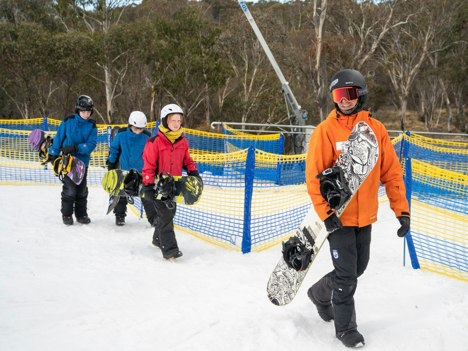 An instructor leads 3 kids, all carrying snowboards, across snow at the bottom of a ski hill