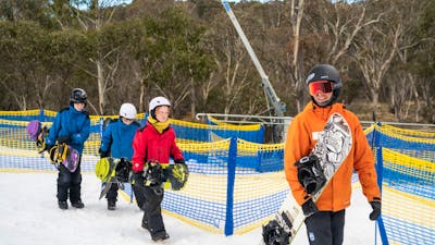 An instructor leads 3 kids, all carrying snowboards, across snow at the bottom of a ski hill
