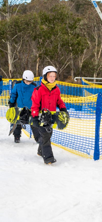 An instructor leads 3 kids, all carrying snowboards, across snow at the bottom of a ski hill