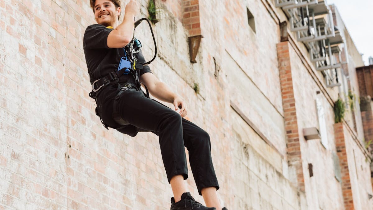 A person in a harness and black clothing smiles while descending a brick wall on a dropline.