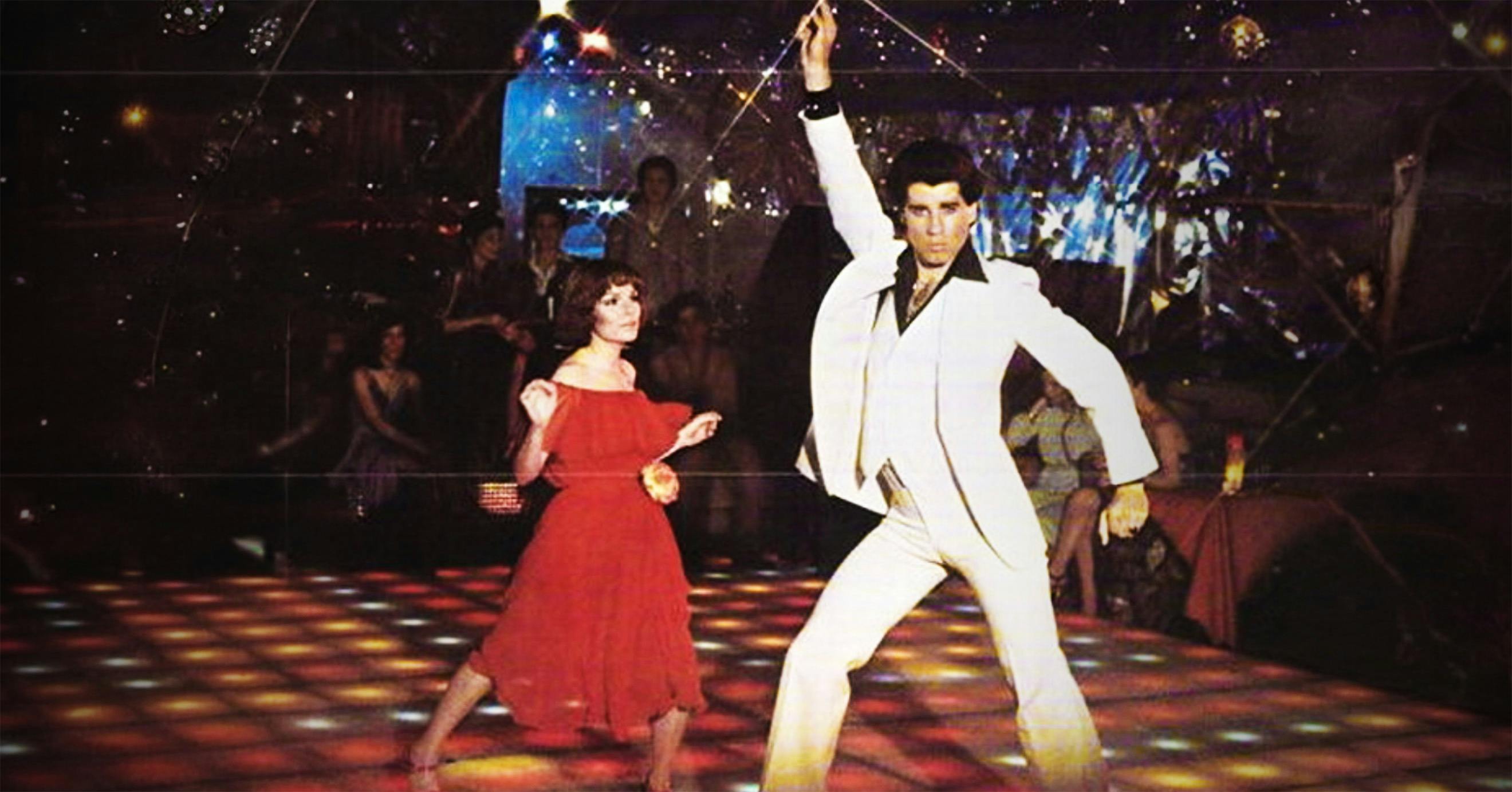 A man in a white suit doing disco moves next to a shorter woman in a red dress dancing