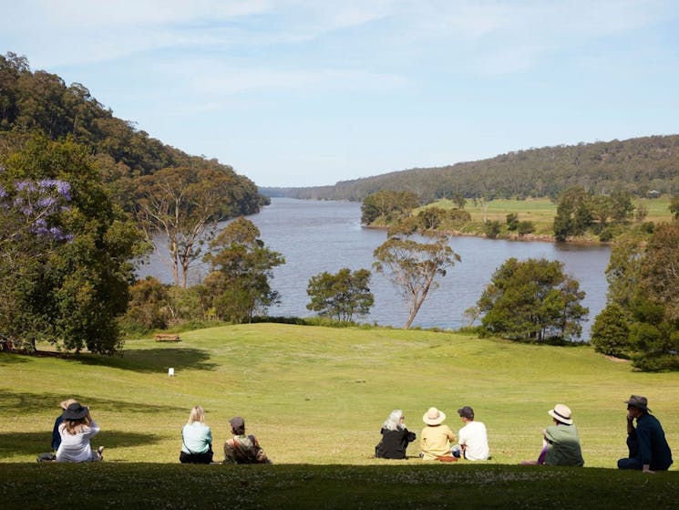 Visitors lounging on the grass lawn at the top of a hill overlooking a bend in the Shoalhaven River