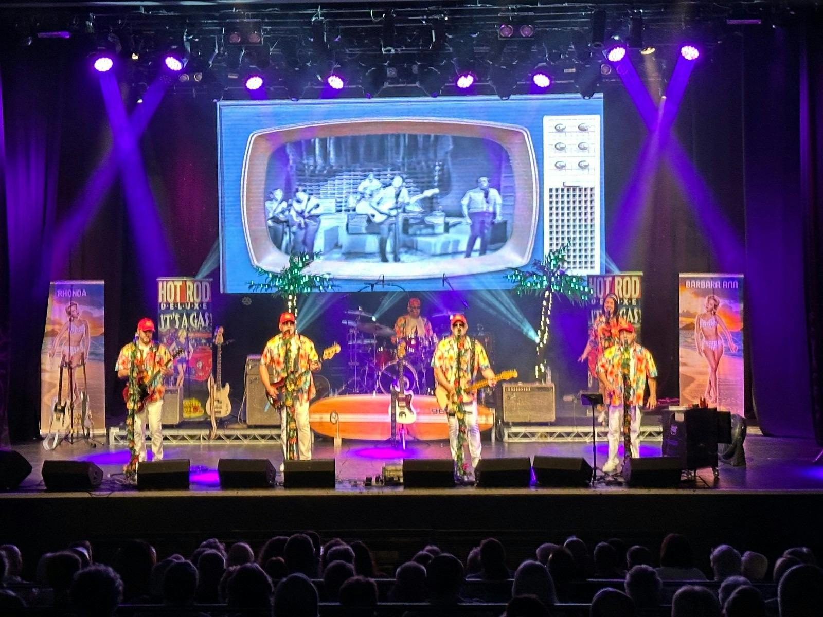Band on stage singing in front of an audience with props on stage behind them