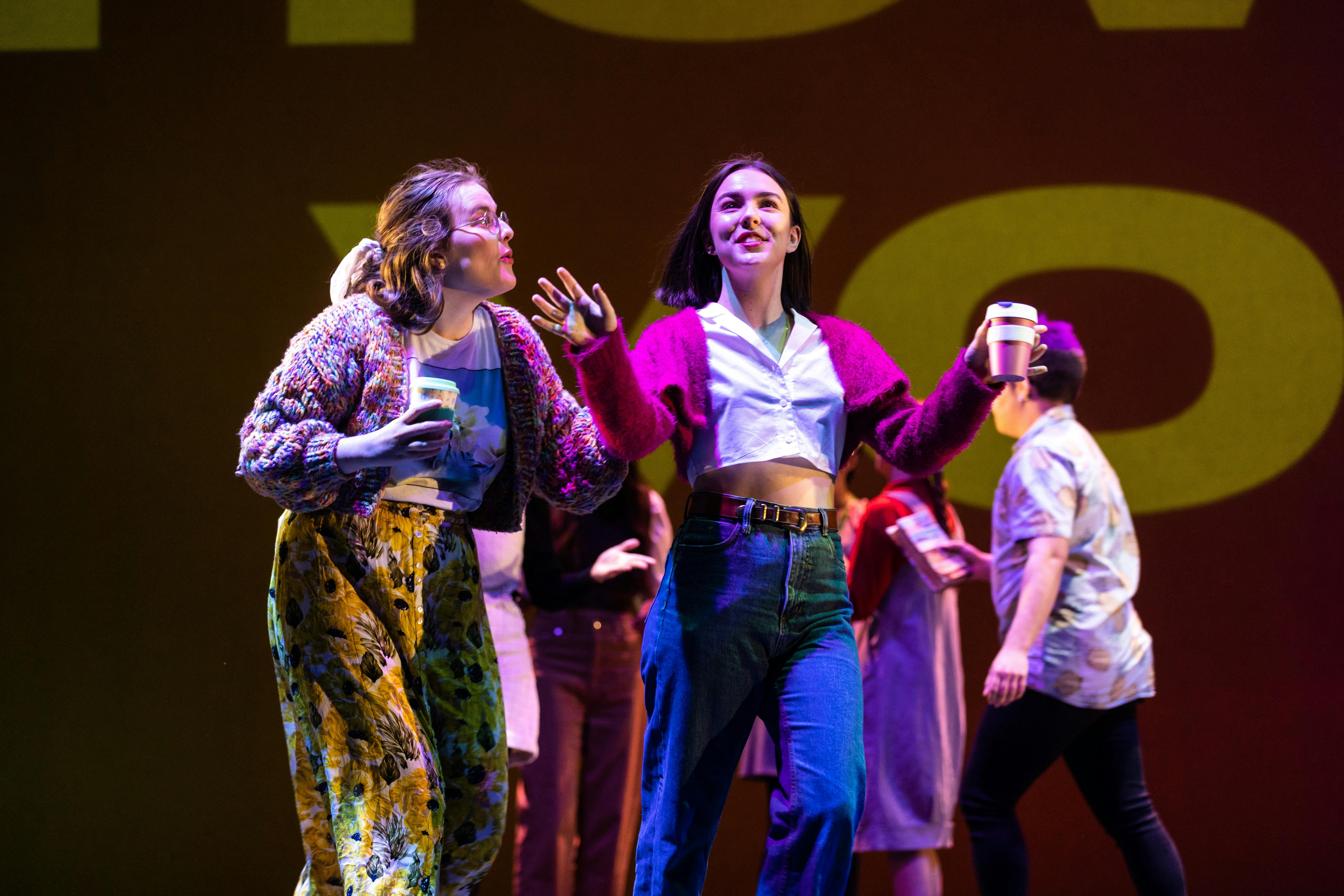 Young actors on stage. A young woman with a coffee cup gestures excitedly and another listens.