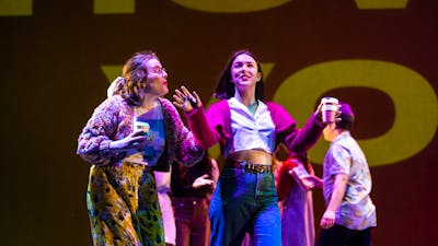 Young actors on stage. A young woman with a coffee cup gestures excitedly and another listens.
