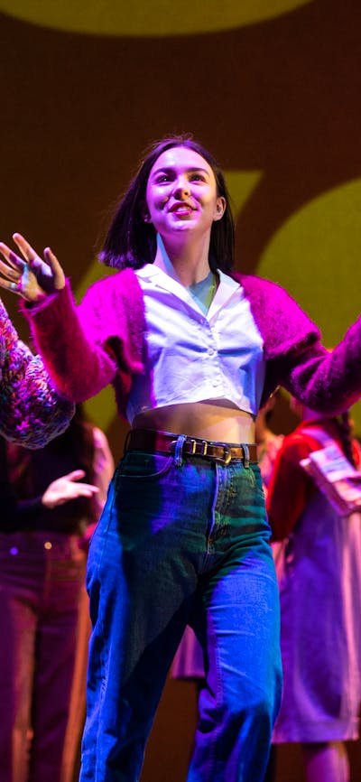 Young actors on stage. A young woman with a coffee cup gestures excitedly and another listens.