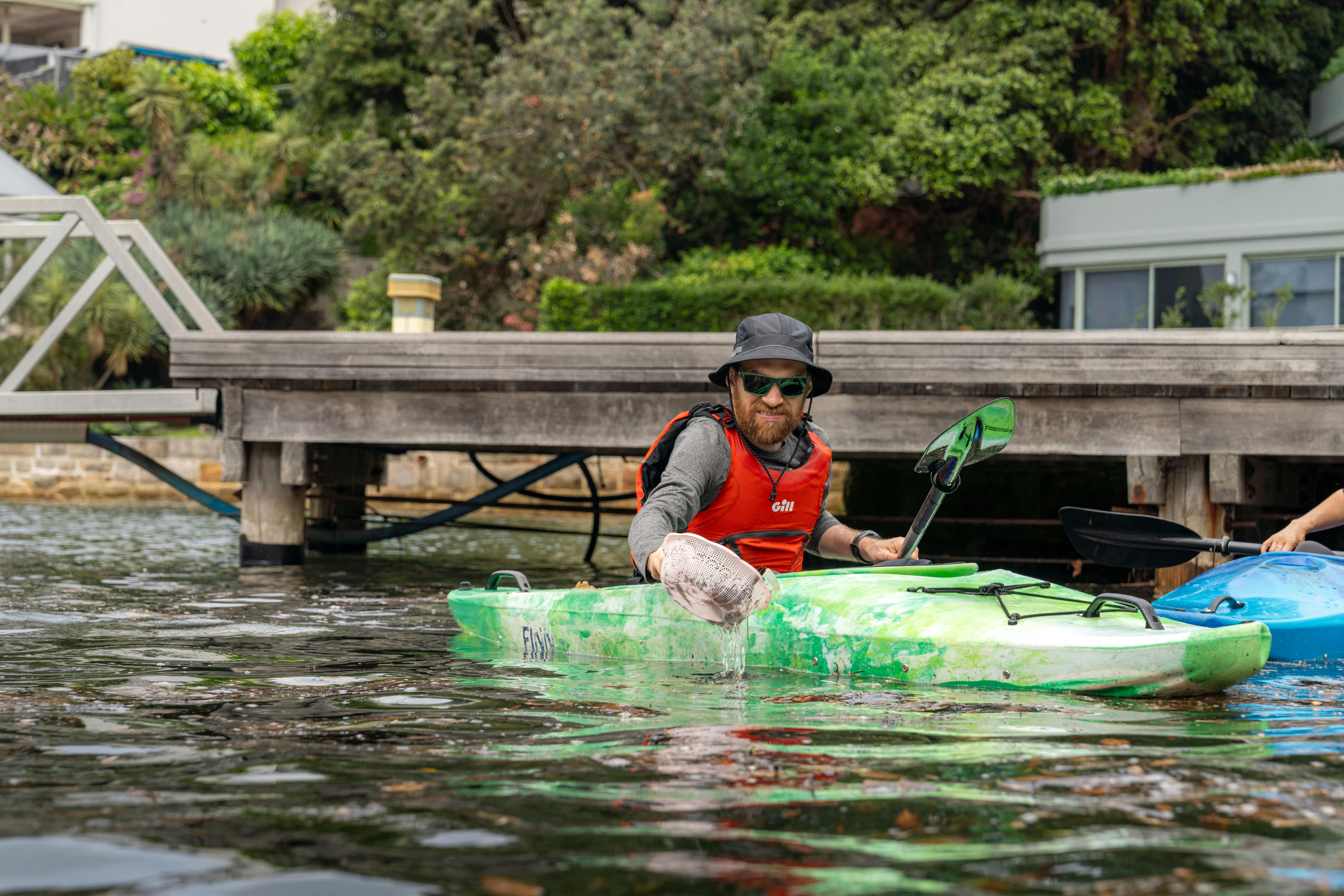 Mann in grünem Kajak reinigt den Hafen von Sydney mit einem Sieb bei einer Aktion von SBK zur Aufräumung von Unternehmen.