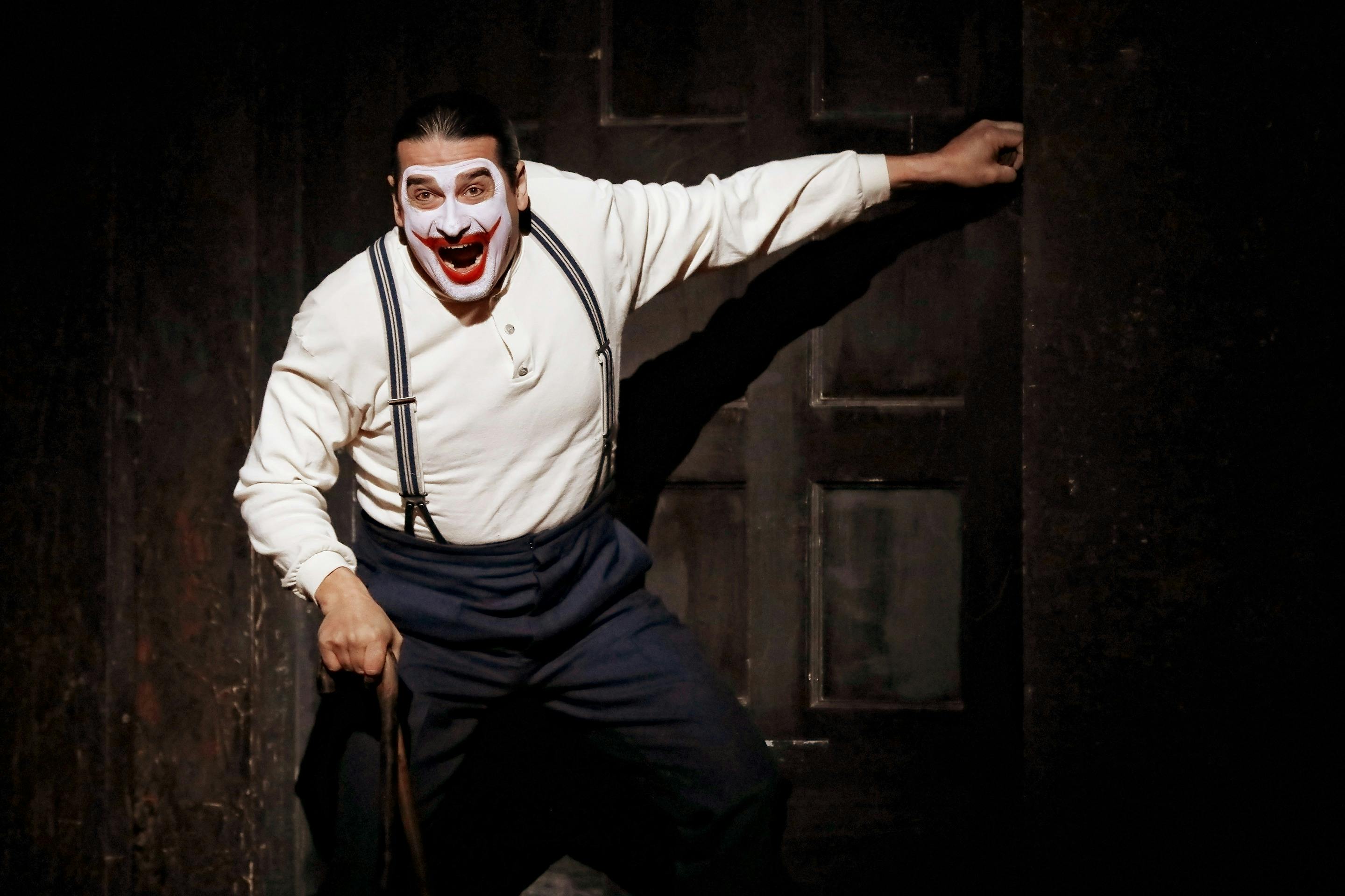 Performer in white clown face paint and suspenders raises arm and sings in dim stage doorway