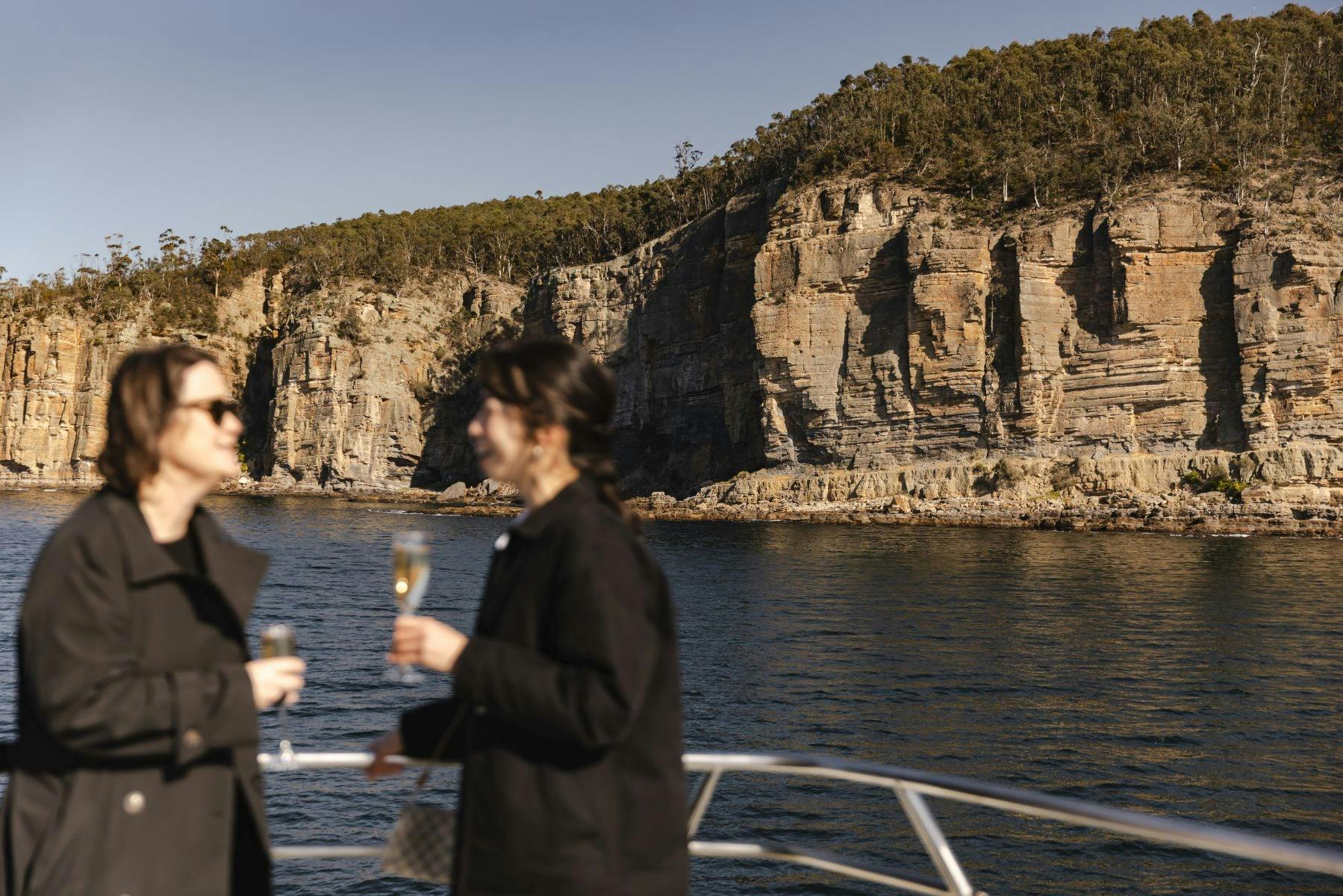 Two women drinking sparkling wine outside on a catamaran, seacliffs in the background