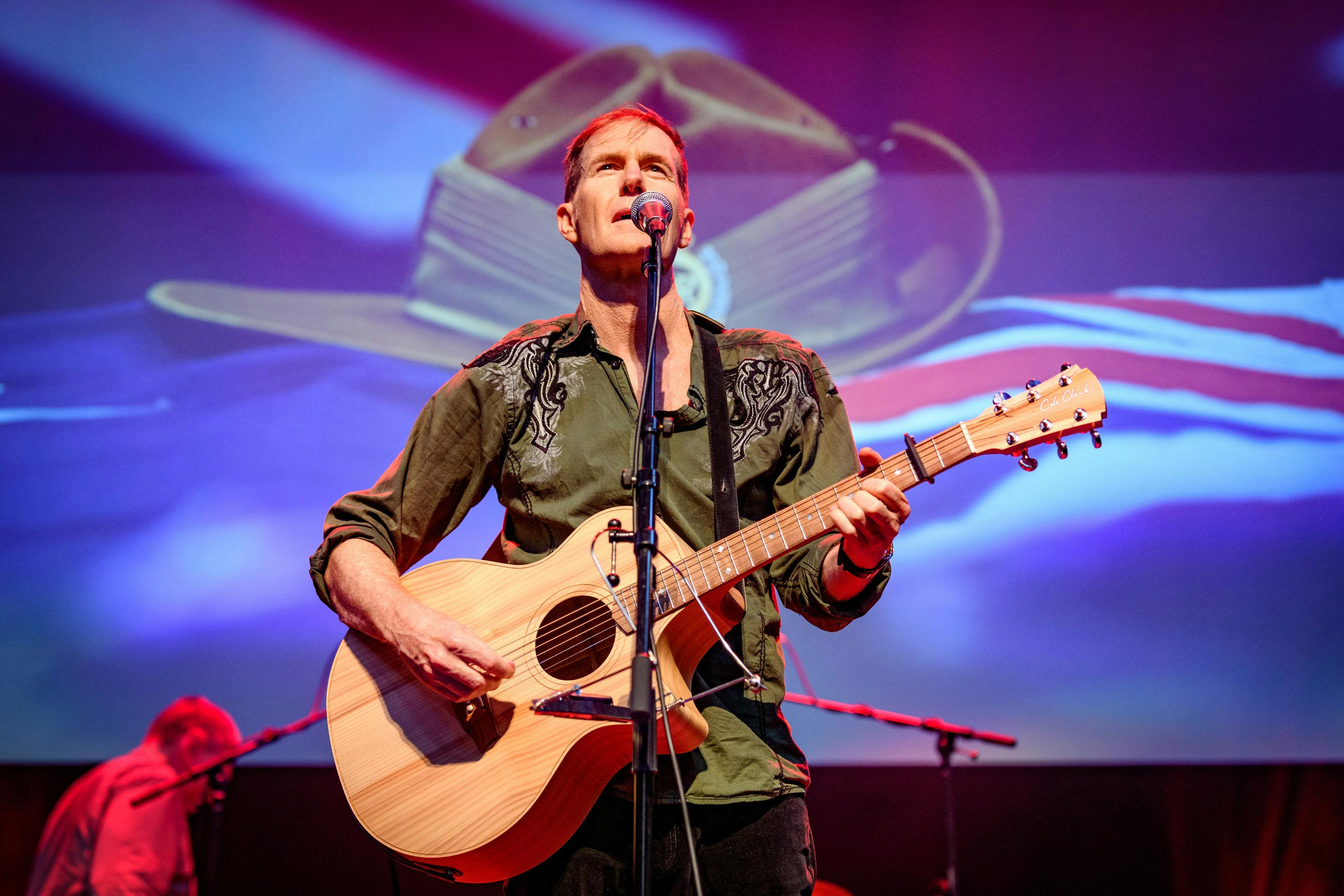 Fred Smith singing and playing an acoustic guitar on stage, in front of a projection of a slouch hat