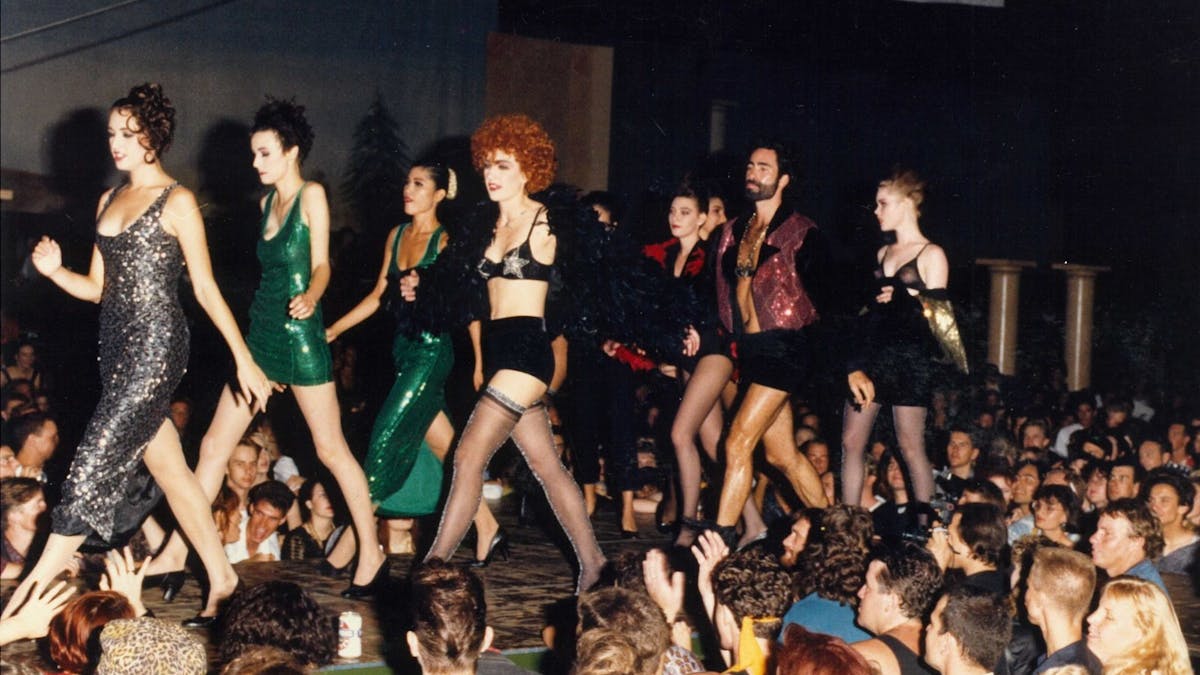 Models walk in a fashion parade wearing  sequin adornments in front of a large crowd