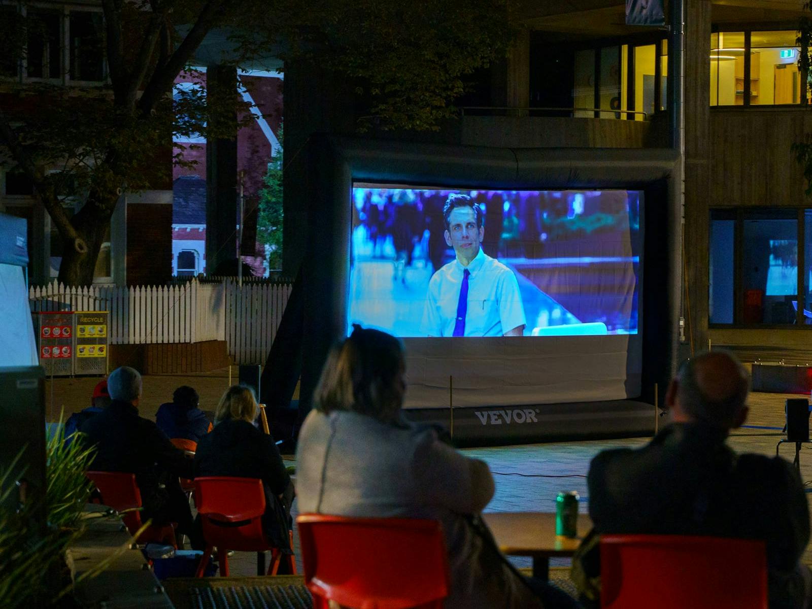 Diners watch a film on the big screen, seated at tables, outdoors.