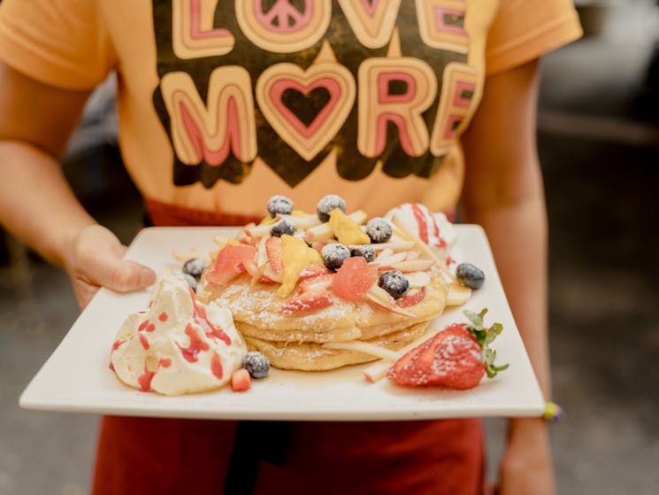 A waitress in a yellow Love More shirt holds a square plate with pancakes and fruit.