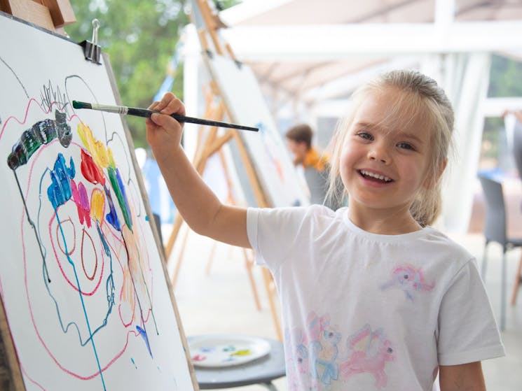 A young blond girl smiling while painting on paper on an easel.