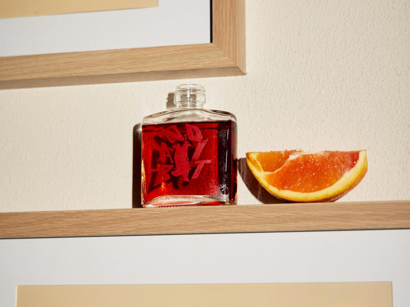 a small square bottle of bright red liquid sits on a wooden picture frame next to a slice of orange