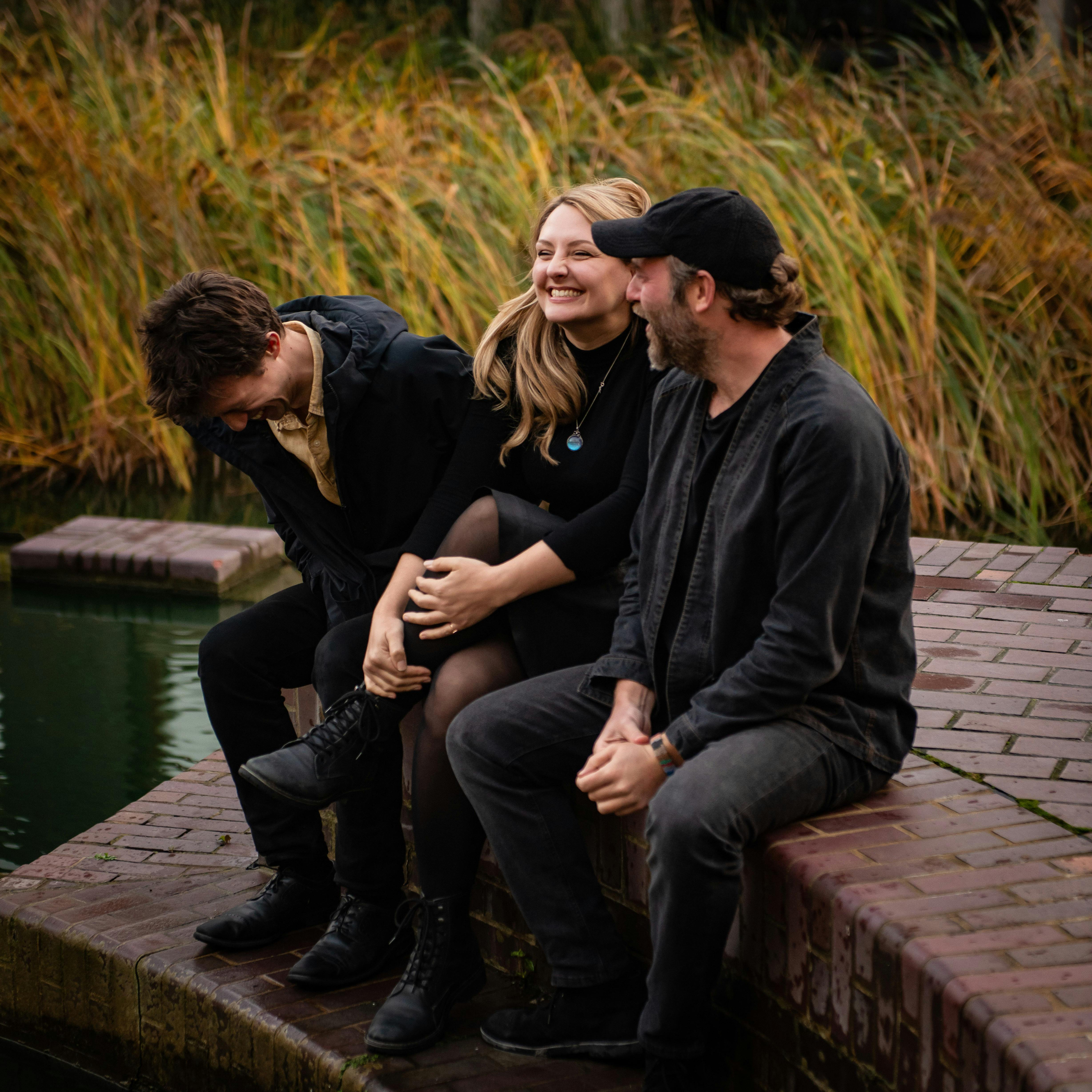 A young man, blonde woman and bearded man laughing together sitting next to a pond
